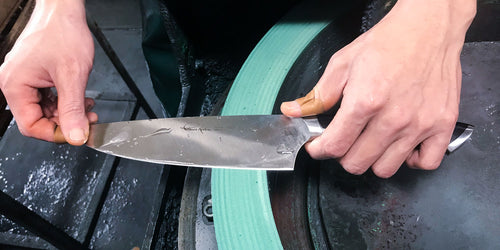 A close up of a bladesmiths hands as the hand sharpen a knife on a green sharpening wheel.
