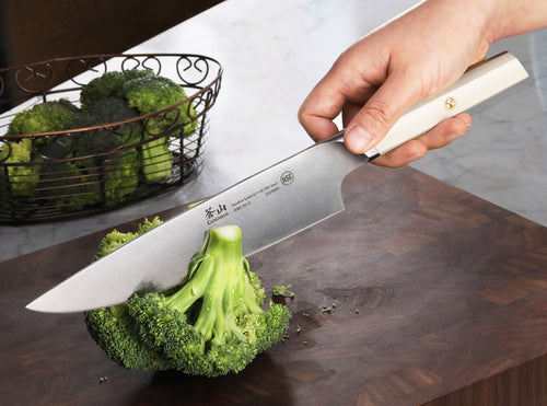 Lifestyle photo of EVEREST series white handle chef's knife cutting through broccoli. 