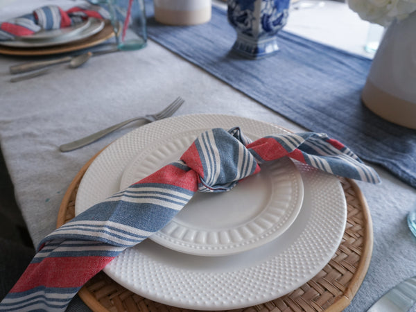 Fourth of July part decor: a red, white, and blue striped napkin, tied in a knot and placed on top of a salad plate, dinner plate, and rattan charger.