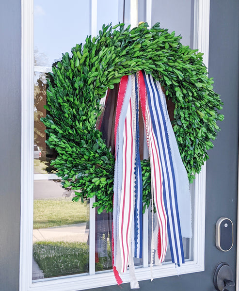 Fourth of july decoriations: boxwood wreath hanging on front door with varieties of red, white, and blue ribbons.