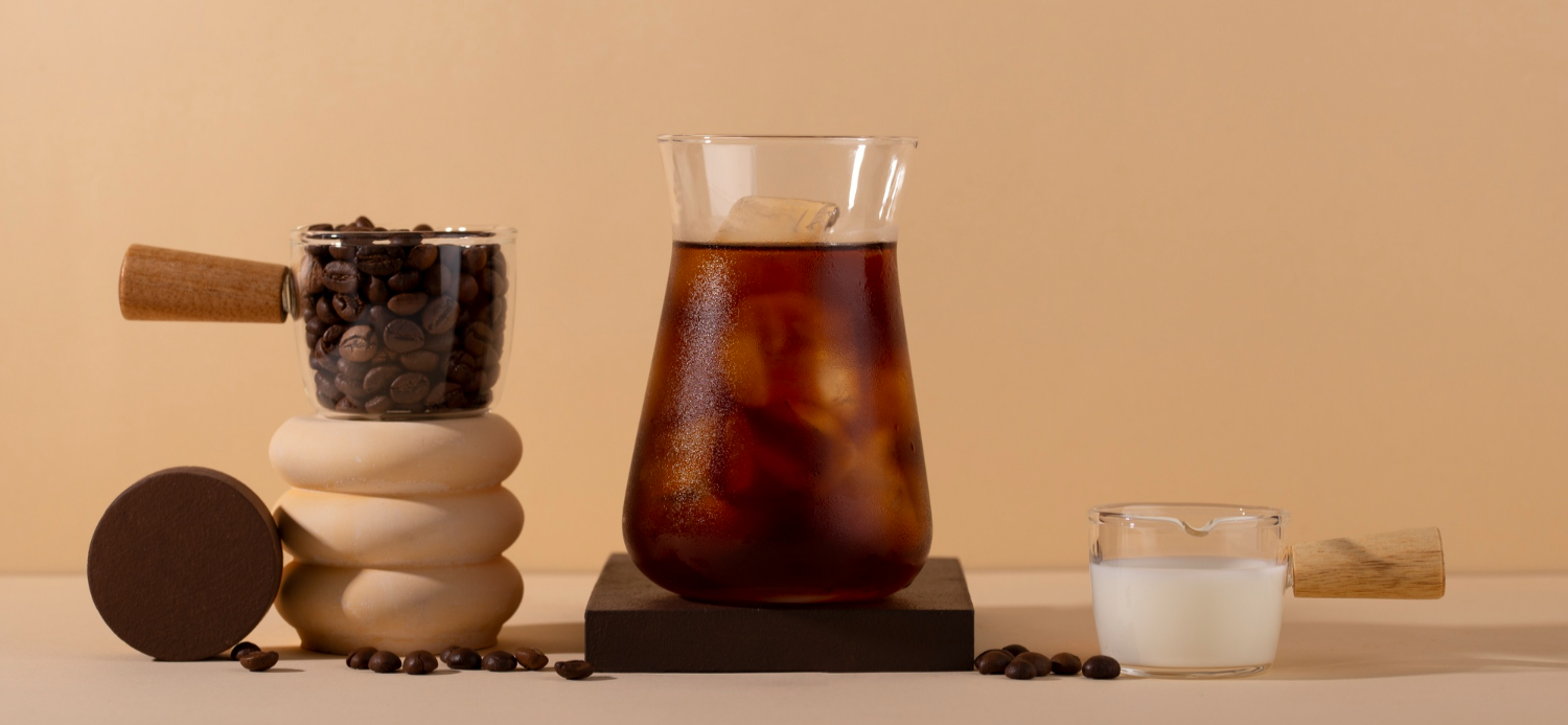 Freshly roasted coffee beans, a vial of cold brew extract, and a glass pitcher of milk arranged together.