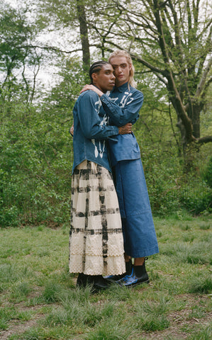 Two models embracing, one wearing a white skirt with black stripes and the other a denim skirt, both models wearing denim tops with white painted designs.