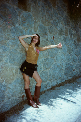 Hippie girl in vintage garb leans against a stone wall waiting for a ride with her thumb extended