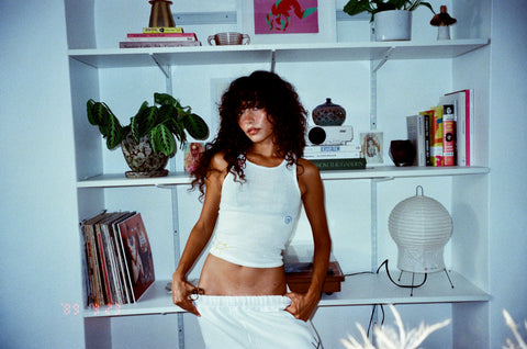 Woman with dark curly hair leans against a book shelf wearing all white leisure clothes that have been embroidered by hand