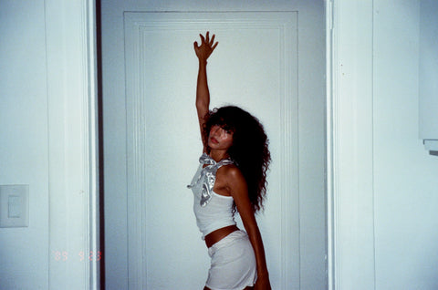 Woman with dark curly hair leans against the hallway walls in a white sleeveless shirt adorned with a bright silver bow
