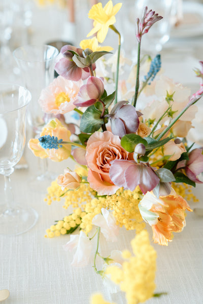 A floral arrangement with peach roses, blue accent flowers, yellow mimosa, daffodils and more spring blooms