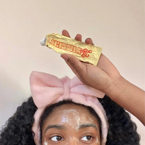 Brightening skin solution is showcased as a person applies a cream from a gold Topicals tube while wearing a pink headband