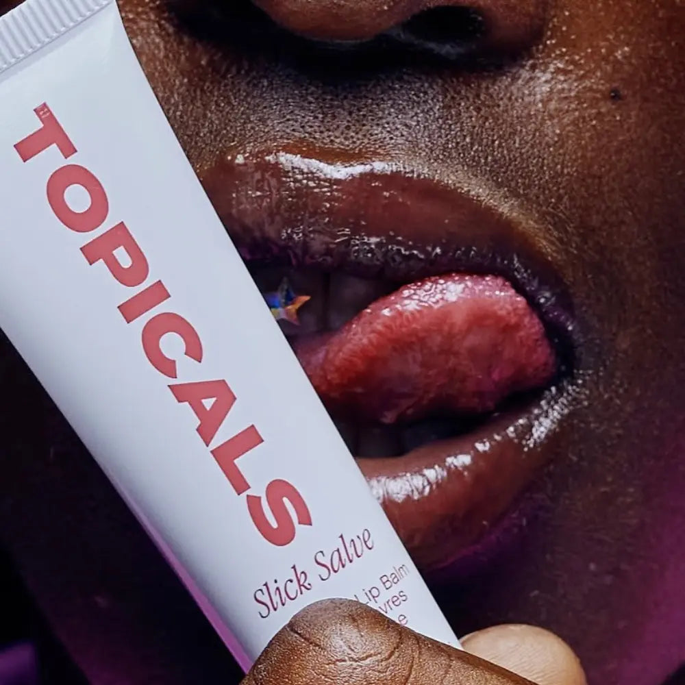 Lip moisturizer held close to glossy lips with a pierced tongue, emphasizing hydration and shine in a bold close-up.