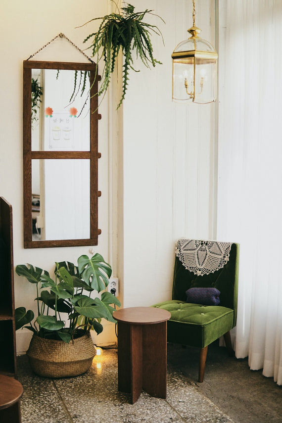 Reading nook with olive green cushion and navy blue back, round dark wood stool at front, basket with monstera on floor, fern in hanging basket, reflected in wood-paneled mirror