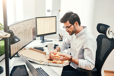 Das Bild zeigt einen Mann, der aus Langerweile vor seinem Computer Pizza isst.
