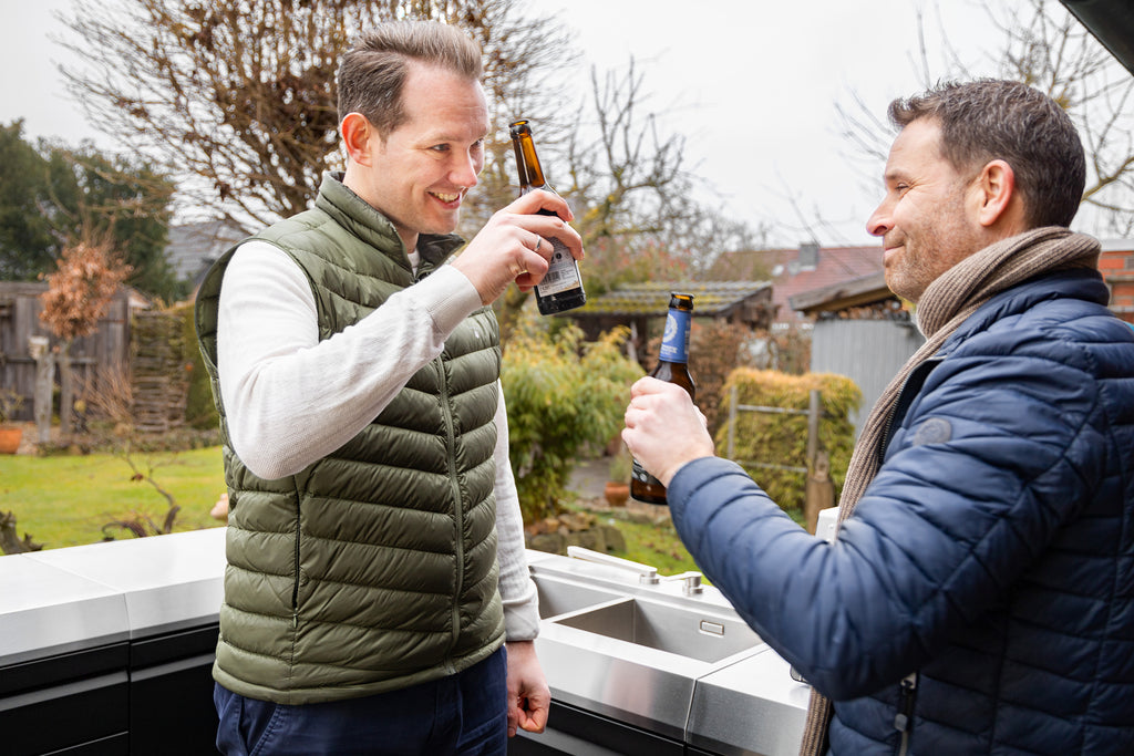Zwei Männer stehen im Garten vor einer Outdoor-Küche und stoßen mit Bierflaschen an