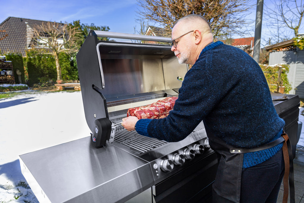 Ein älterer Mann steht vor dem Grill seiner Outdoor-Küche und setzt eine Rotisserie mit Fleisch ein