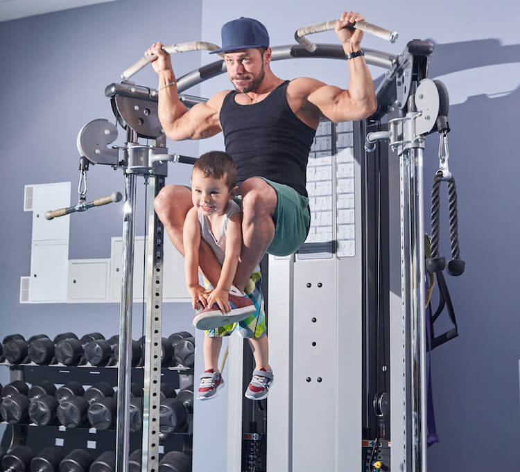 father excercising with his young son in a gym, holding him with his legs while he's working on his arms area