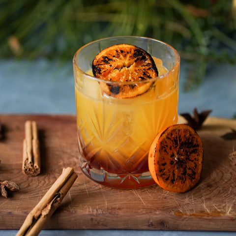 orange and deep red two tone cocktail in a rocks glass garnished with charred clementine against a festive backdrop of greenery