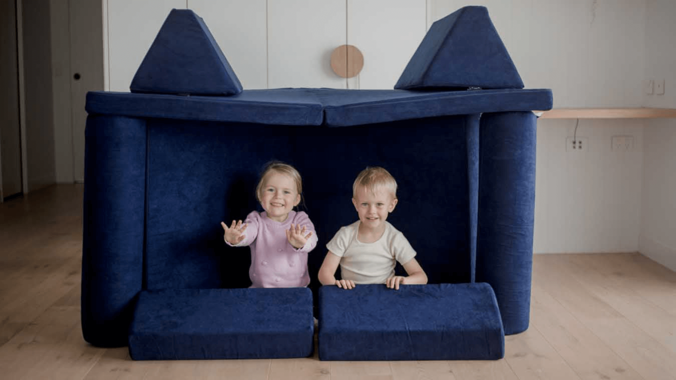 Two toddlers are sitting on a Midnight Blue Funsquare® Kids Play Couch Set that is set up like a cubby. They are both looking at the camera and the child on the left has their hands outstretched.