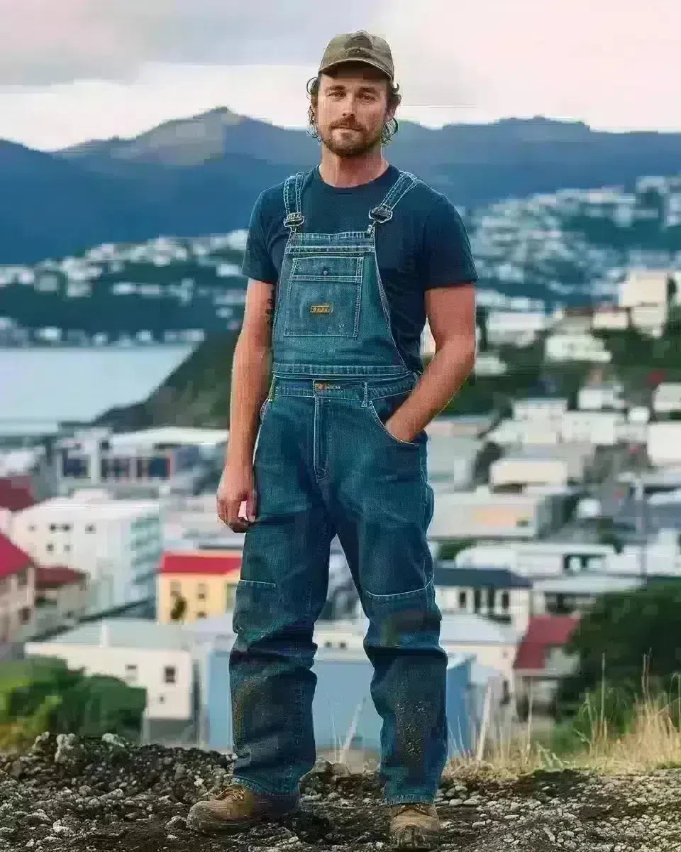 Man in denim overalls stands before Wellington's skyline, showcasing durability and utility. Late Winter  season.