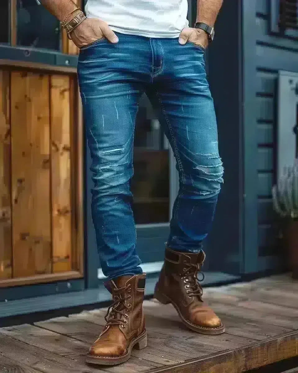 Man in indigo raw denim jeans, white tee, chestnut boots, outdoor urban setting. Spring season.