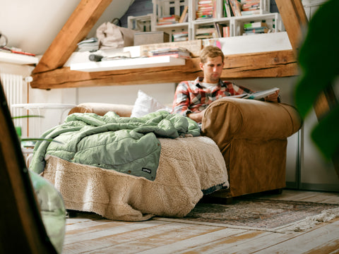 Blick in ein Wohnzimmer, wo ein Mann auf der Couch sitzt und ein Buch liest, eingewickelt in eine salbengrüne Kuscheldecke