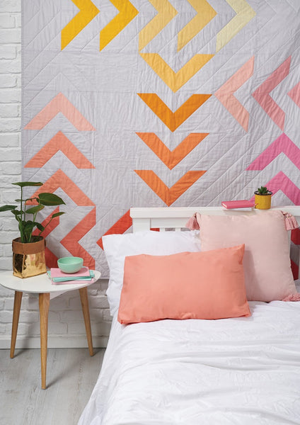 Dayglow quilt pattern by Penny Spool Quilts - quilt in pink, peach and yellow on grey background, hanging behind a bed with peach and white bedding