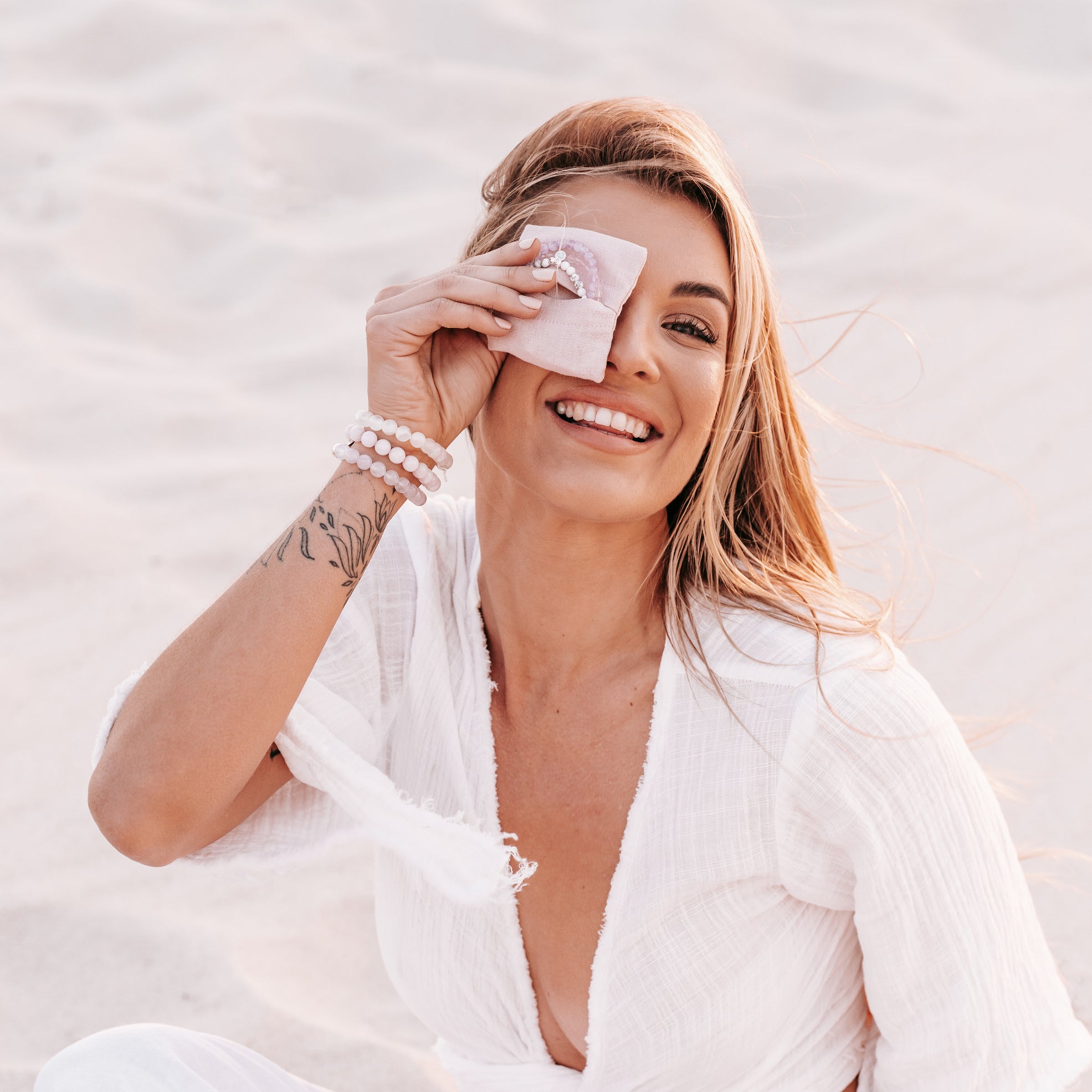 Happy blonde smiling model at the beach holding linen pouch over her eye wearing crystal 925 bracelets