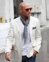 Man walking with one hand in his pocket, wearing a white blazer, tshirt, pocket square, small scarf, and sunglasses.