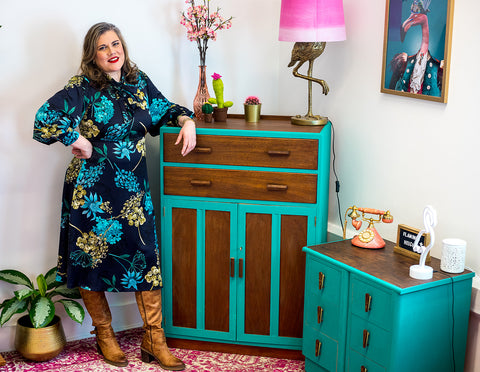 Image of Corinne stood in front of a chest of drawers wearing a turquoise and dark blue dress