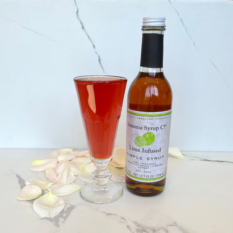 Cranberry lime mimosa in champagne glass next to a bottle of Sonoma Syrup Co. Lime Syrup with flower petals in background