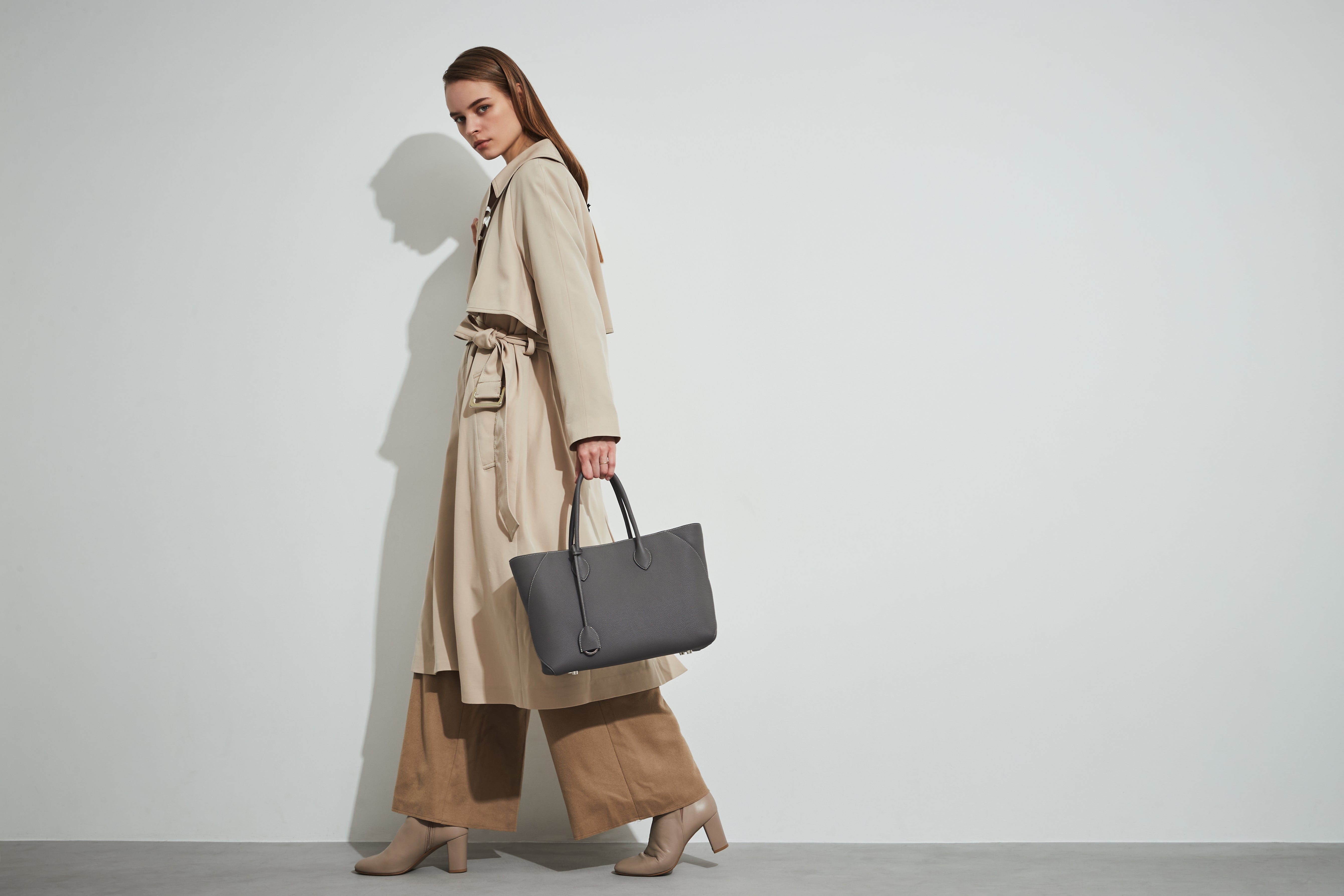 Elegantna ženska v trenčkotu nosi torbo Mia Tote Bag Medium blagovne znamke BONAVENTURA, ki je dovolj prostorna za računalnik Macbook in je idealna za vsakdanjo uporabo.