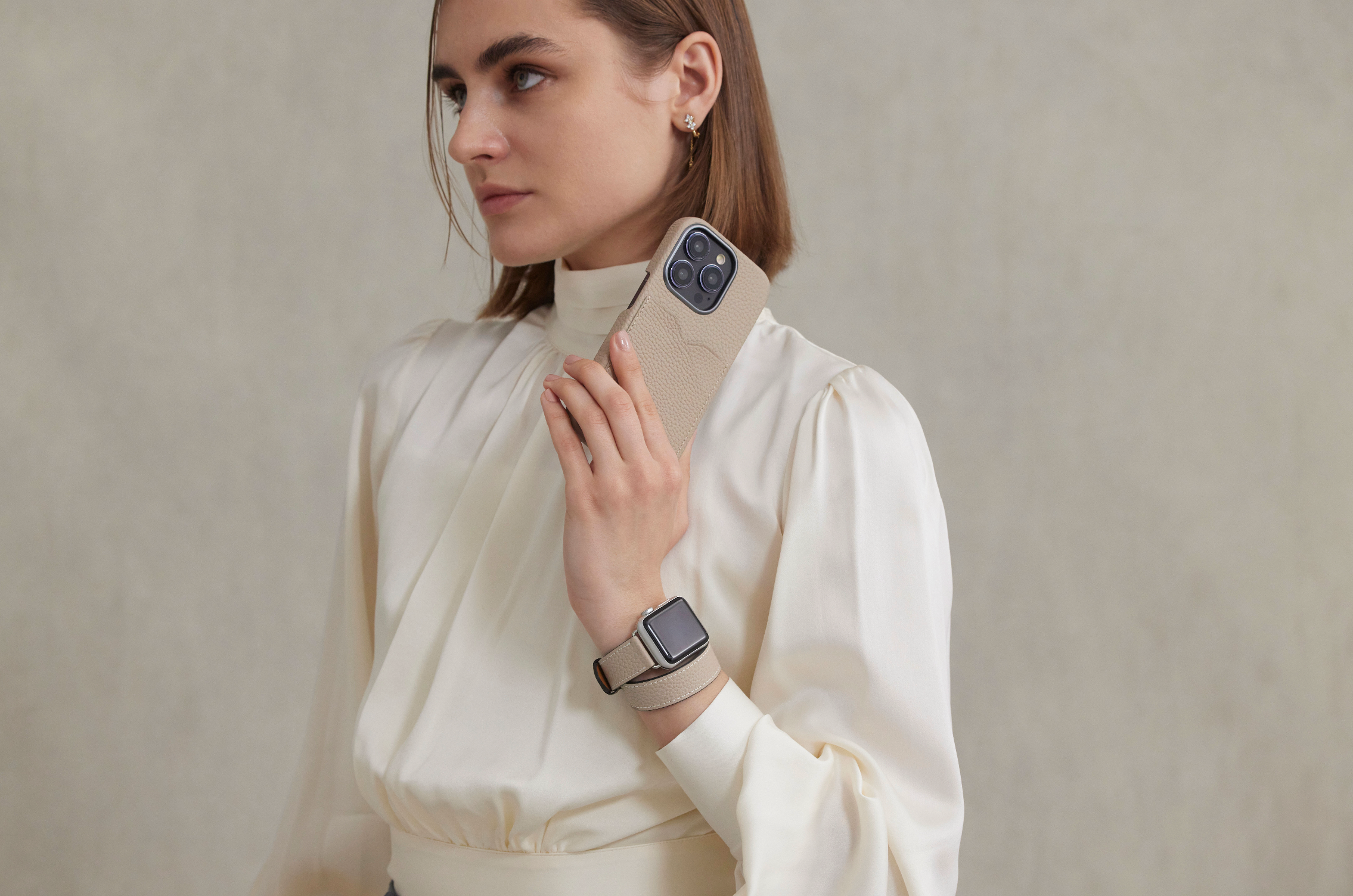 Stilīgi ģērbta sieviete valkā augstas kvalitātes ādas siksniņu savam Apple Watch un elegantu ādas vāciņu savam iPhone 15.