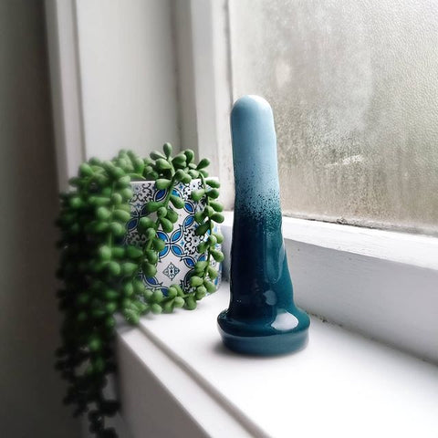A 4 inch classic ceramic dildo in a dark green to light blue gradient pattern stands on a windowsill. A green plant in a blue and white patterned pot stands in the background.