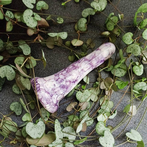 A 4 inch classic ceramic dildo in a purple bubble pattern lies on a grey surface. Tendrils from a chain of hearts plant flow around the dildo.
