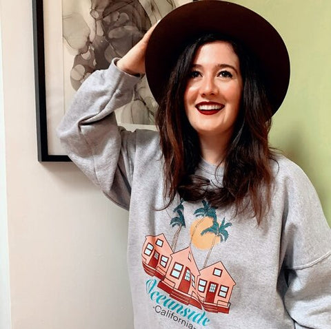 A femme presenting person stands in front of a green and white wall with a black and white abstract artwork in a black frame. They are wearing a black sun hat over their dark brown hair and are wearing a grey graphic printed sweatshirt.