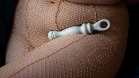 A white ceramic sex toy is nestled in the curve of a person's hip and thigh. The person wears tan-toned fishnet stockings.