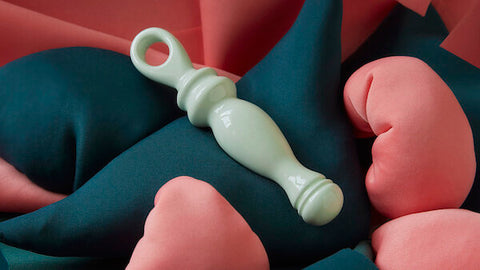 A mint coloured ceramic sex toy lies on a bed of small cushions which are teal and coral coloured.