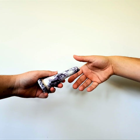 A hand holds a 4 inch ceramic dildo in a purple bubble pattern, while another hand reaches for it, against a white background