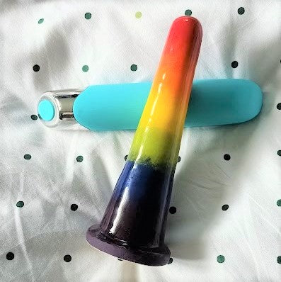 A 6 inch classic ceramic dildo in a rainbow gradient pattern is propped up on a teal coloured large bullet vibrator. Both toys lie on a white sheet with dark green dots.