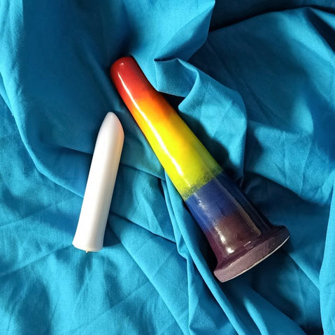 A 6 inch classic ceramic dildo in a rainbow gradient pattern and a white bullet vibrator lie on a rumpled blue sheet