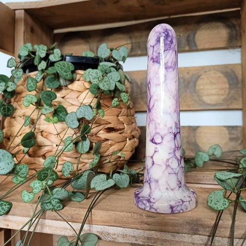 A 5 inch classic ceramic dildo in a purple and pink bubble pattern stands in a wooden crate. Tendrils from a chain of hearts plant flow from a wicker pot around the dildo.