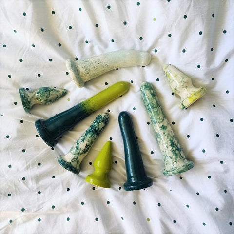 A selection of green patterned ceramic sex toys lie together on a white sheet with green dots.