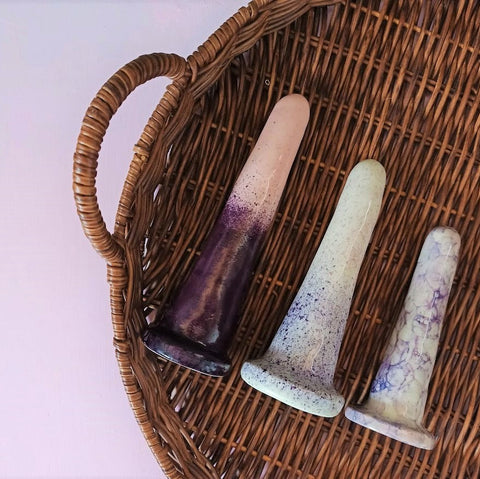 3 classic ceramic dildos in 6, 5 and 4 inch sizes lie on a wicker stand on a pink surface. The patterns on the dildos are all purple variations.