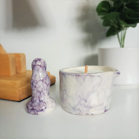 A small ceramic butt plug and a matching ceramic massage candle in a dark purple bubble pattern stand on a white bedside table. A wooden lamp and a dark green plant in a white pot are visible in the background.