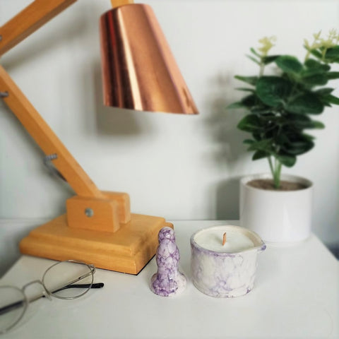 A small ceramic butt plug and a matching ceramic massage candle in a dark purple bubble pattern stand on a white bedside table. A pair of wire-rimmed glasses lay to the left, and a wooden lamp with a copper shade and a dark green plant in a white pot are visible in the background.