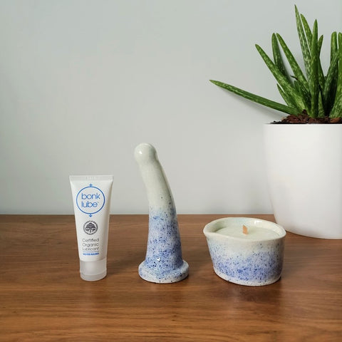A tube of organic lubricant, a 6 inch curved ceramic dildo and a matching massage candle in a blue speckle pattern stand in a row on a wooden table. An aloe vera plant in a white pot is visible in the background to the right.