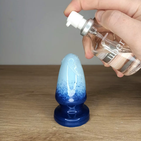 A large butt plug in a dark blue to light blue gradient pattern stands on a wooden table. A hand holds a bottle of Uberlube lubricant above the butt plug and is squirting lubricant onto it.