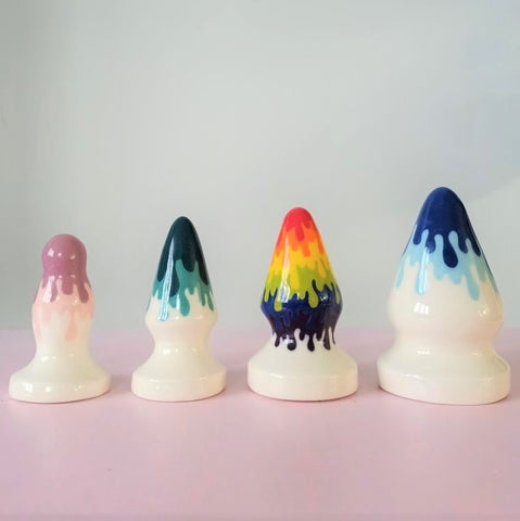 4 ceramic butt plugs of increasingly large size stand in a row on a pink table, from smallest on the left to largest on the right. They all have a drip pattern from left to right; of purples, greens, rainbow and blues.