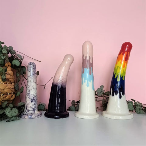 4 colourful ceramic dildos of various patterns and sizes stand on a white table in front of a pink background. Tendrils from a chain of hearts plant flow from a wicker pot on the left around the dildos.