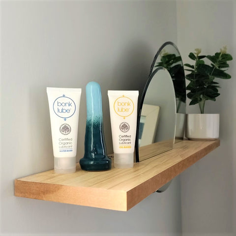 Two tubes of Bonk organic lubricant stand either side of a 4 inch classic ceramic dildo in a dark green to light blue gradient pattern. The items stand at the end of a wooden shelf on a white wall, with circular mirrors inserted into the shelf. A pot plant stands at the other end of the shelf.
