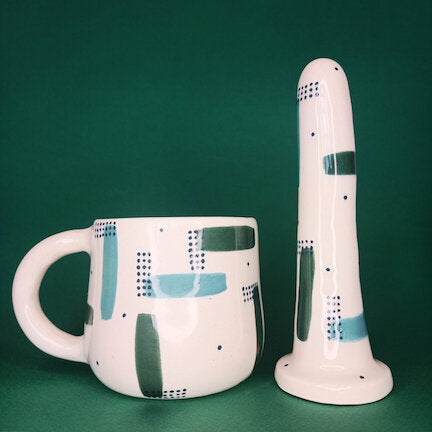 A 5 inch classic ceramic dildo and a matching mug with green and teal strokes and dark green dots stand in a dark green photobooth. 
