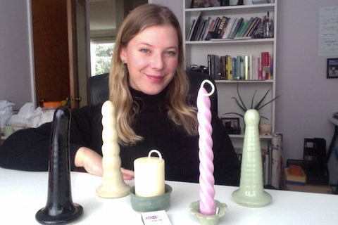 A femme presenting person sits in front of a white table with an assortment of ceramic sex toys and candles. They have medium length dark blonde hair and are wearing a black turtle neck sweater.
