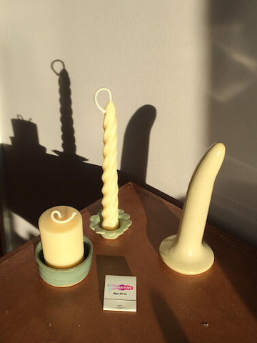 A cream coloured ceramic dildo stands on a wooden table next to two candles and a business card.